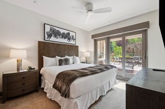 A master bedroom in The Charter F2305 featuring a large bed with a dark wood headboard, white linens, a faux fur throw, a nightstand with a lamp, and glass doors leading to a private patio.