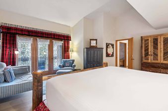 A cozy bedroom in The Charter E430 features a large bed, a daybed, and a blue armchair, with glass doors leading to a balcony, framed by plaid curtains.