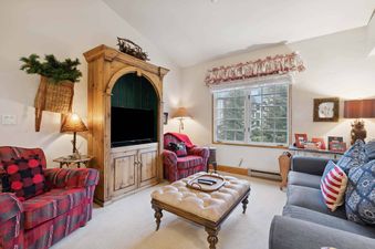 A cozy lounge area in The Charter E4305 features plaid armchairs, a gray sofa, a large wooden entertainment center with a television, and a window with a floral valance.