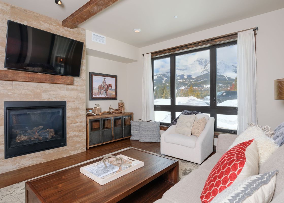 A cozy and modern living room with a fireplace, a large TV, comfortable seating, and a window showcasing a stunning mountain view.