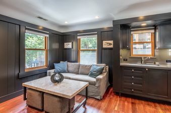 The living area of Cedar Crest Woodpecker Cottage 7 features dark wood paneling, a gray sofa with blue pillows, a coffee table with ottomans, and a compact kitchenette with a sink and dark cabinets.