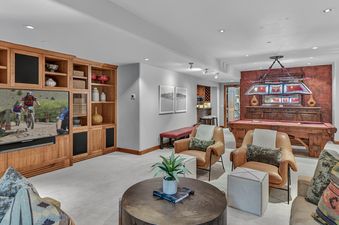 The Timber Haven features a spacious games room with a large wooden entertainment center, a flat-screen TV displaying a mountain biking scene, comfortable seating, and a classic billiards table under an ornate chandelier.