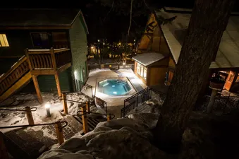 A beautifully lit outdoor hot tub at night, surrounded by snow and two cozy cabins, inviting relaxation in the winter ambiance.