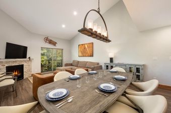 The dining area in Vasquez Creek Townhomes 8 features a large wooden table with place settings, positioned beneath a stylish chandelier, leading into a cozy living space with a fireplace.
