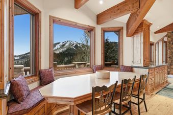 The dining area at Summit View Lodge at Deer Valley features a large wooden table, chairs with tree-silhouette designs, and built-in window seating, all set against a backdrop of snow-capped mountains and lush forests.