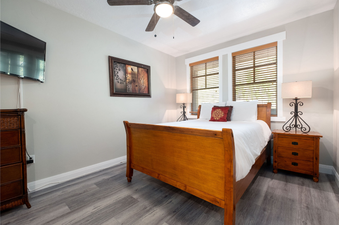 A cozy bedroom in Town Pointe C106 features a wooden sleigh bed with white bedding, two bedside tables with lamps, a flat-screen TV, and windows with blinds.