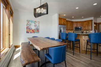 A modern dining area at EagleRidge Townhomes 1461 with a live-edge wooden table, blue chairs, and a geometric chandelier, adjacent to a kitchen with wooden cabinets and stainless steel appliances.