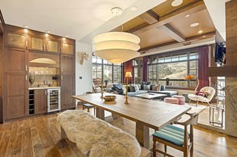 An expansive open-concept living and dining area at Guardsman Overlook, featuring a large wooden dining table with a fur-covered bench, a tiered pendant light, a blue sectional sofa, and expansive windows offering mountain views.