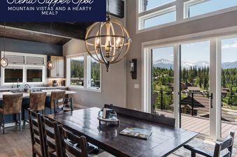 The dining room and kitchen of Peak View Paradise feature a long wooden table and an open kitchen with bar seating, all bathed in natural light from large windows offering expansive mountain views.