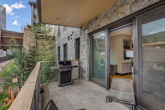 A private balcony at One Snowmass East 304 featuring a BBQ grill, stone facade, and sliding glass doors leading to the interior, with lush greenery and mountain views in the background.