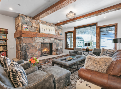 Grand Lodge 3 Bedroom Deluxe 304 living room with a stone fireplace and leather sofas and a large window that looks out onto a snow-covered mountain.