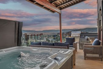 A luxurious outdoor rooftop terrace at sunset, featuring a private hot tub, a comfortable lounge area with dark blue cushions, and a stainless steel BBQ grill, all overlooking a city and distant mountains.