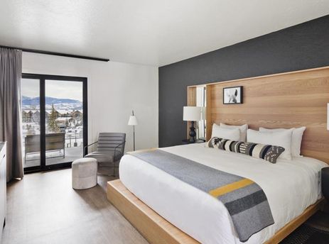A modern bedroom with a king bed, grey armchair, and sliding doors leading to a balcony with snowy mountain views.