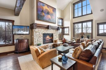 A spacious and luxurious living room at Silver Star 47 featuring a stone fireplace, high ceilings, large windows with winter views, and comfortable leather seating.