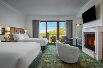 Two queen beds with white linens and brown headboards face a fireplace with a mounted television above it, and a glass door leads to a balcony with a mountain view.