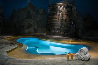 A serene indoor pool at Phillips Ridge with glowing blue water, a waterfall over a stone wall, and two tropical drinks with neatly stacked white towels nearby.