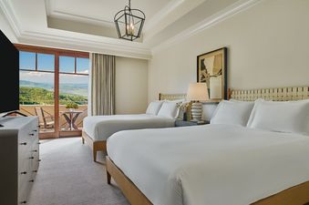 Two queen beds with white linens and light wood frames are shown in a room with light-colored walls and a view of green mountains from a large glass door.