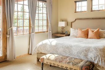 A cozy bedroom at Docs Holiday with a large bed, patterned duvet, orange pillows, wooden side table, and windows overlooking the outdoors.