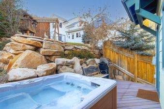An outdoor hot tub and grill area with a wooden deck, surrounded by a rock wall and trees, with Woodside Manor visible in the background.