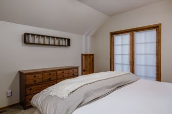 A cozy bedroom at Southfork Retreat featuring a bed with white and gray bedding, a wooden dresser, a unique wall decoration, and wooden-framed doors with white blinds.