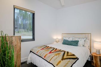 The bedroom in Moonlight Basin 12 Lake Cabin has a white bed with a gold headboard, white bedding and a colorful throw blanket. There is a window with a view of trees outside and a wooden dresser on the left.