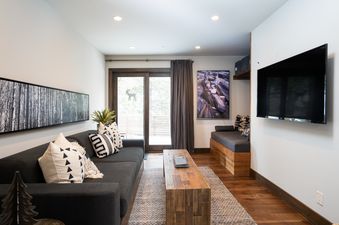 A modern living room at the West Shore Hideaway features a dark gray sofa with geometric pillows, a wooden coffee table, a panoramic forest artwork, a wall-mounted TV, and glass patio doors with an outdoor view.