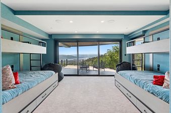 A modern bunk room with two bunk beds, blue bedding, and a balcony overlooking mountains and a lake.
