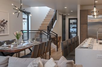 The dining area in the Principal House features a large wooden table set for a meal, a modern chandelier, and an adjacent kitchen island with bar seating.