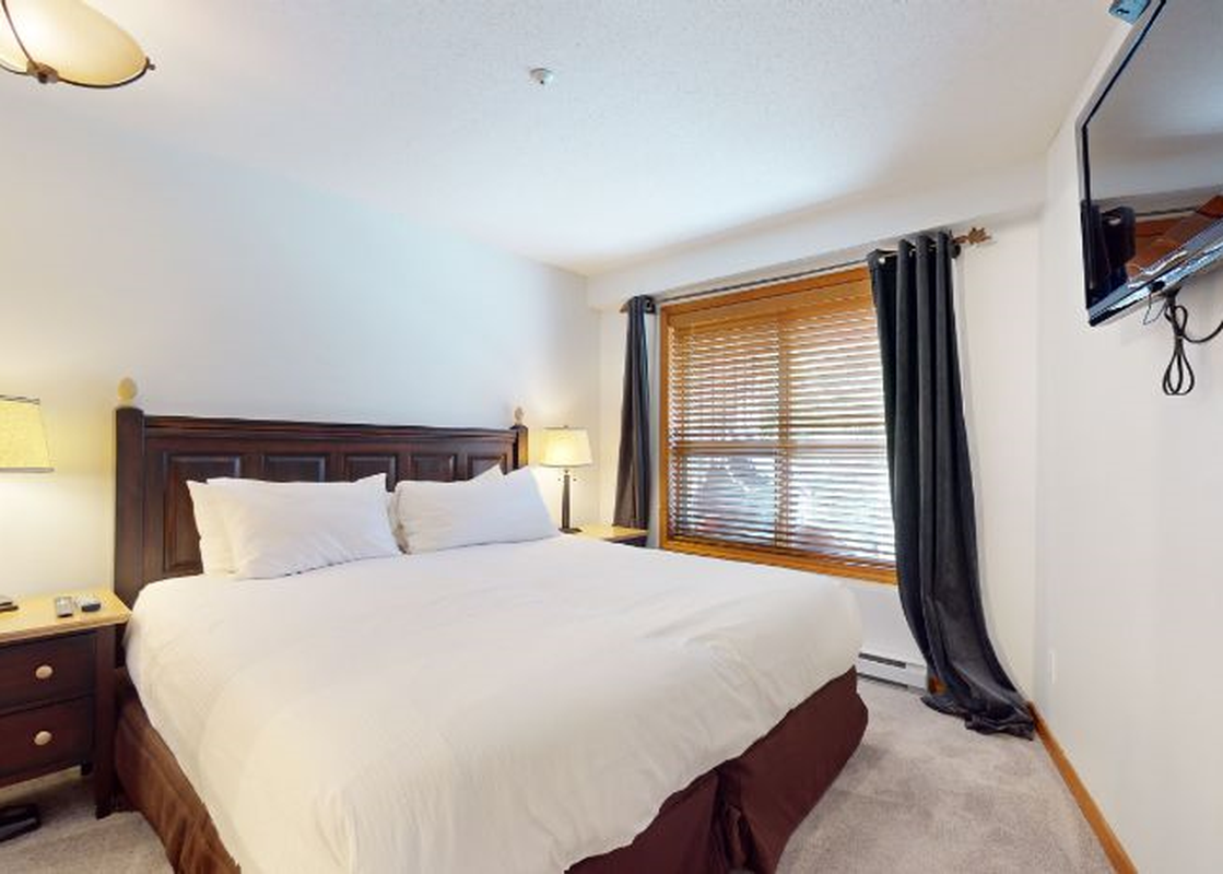 The bedroom in Aspens on Blackcomb Unit 332 features a large bed with a dark wooden headboard, flanked by two nightstands with lamps, and a window with blinds and dark curtains.