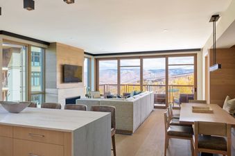 A spacious, open-concept living and dining area in the Cirque Two Bedroom Den Residence, featuring modern furnishings, a fireplace, and large windows with mountain and ski lift views.