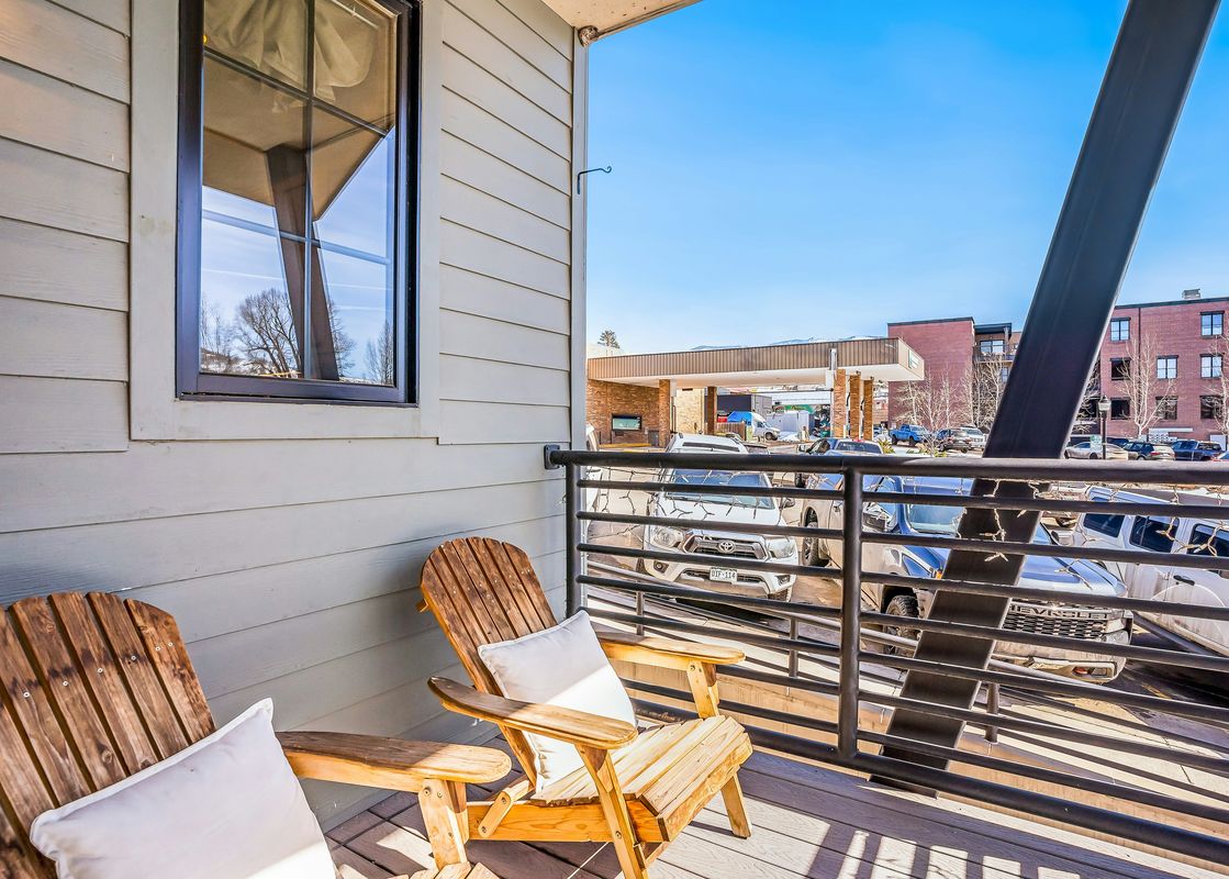 A wooden balcony with two chairs and a view of a parking lot and a building in the distance.