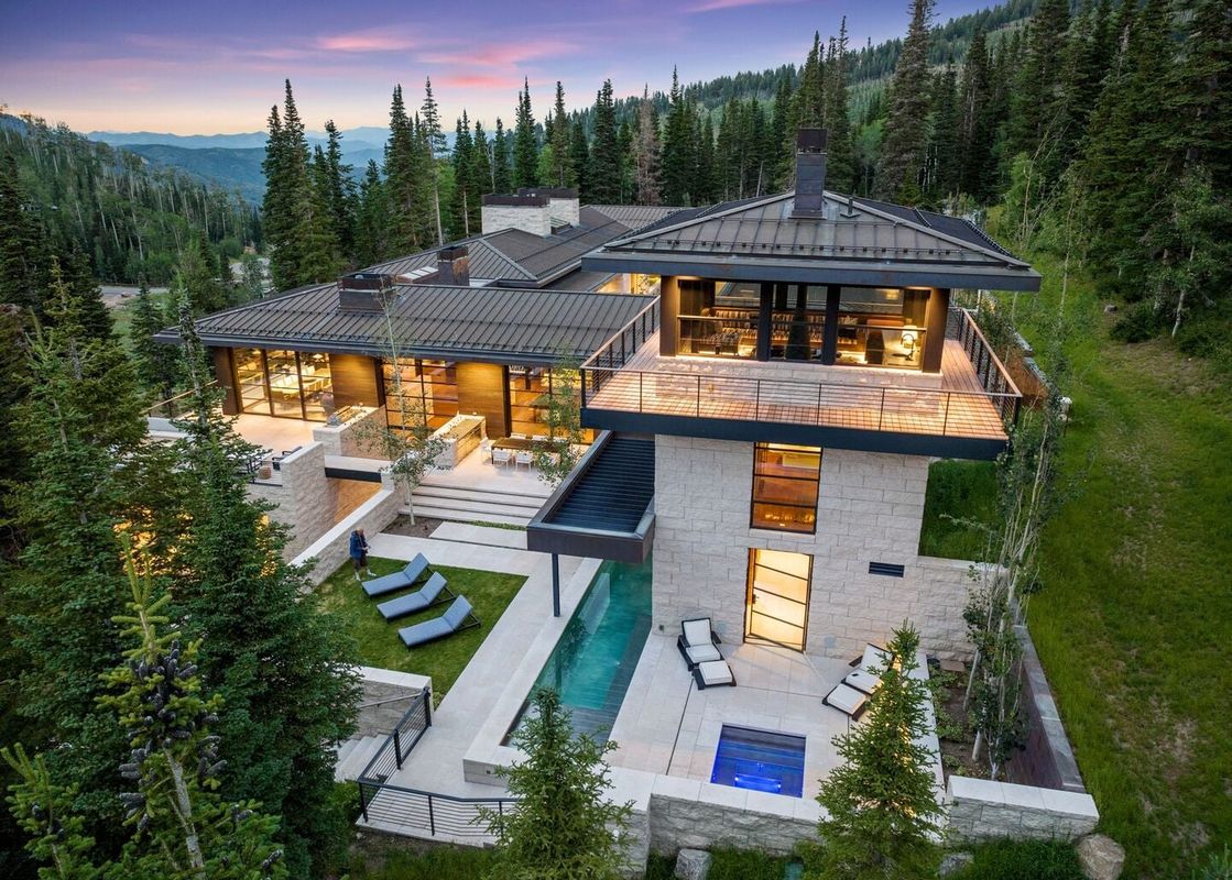 An aerial view of the Monitors Rest, a modern mountain home with multiple levels, outdoor living spaces, a swimming pool, and an elevated balcony, surrounded by a dense forest under a twilight sky.