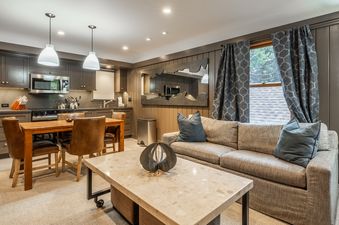 A modern open-concept kitchen and living area with gray cabinetry, stainless steel appliances, a wooden dining table, a gray sofa with blue pillows, and a unique framed mirror.