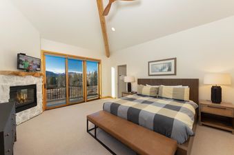 A spacious bedroom at Moosehead Lodge, featuring a king-size bed with plaid bedding, a stone fireplace, and large glass doors opening to a balcony with mountain views.