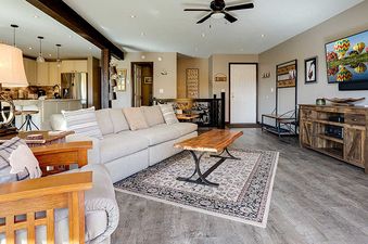 A bright and spacious living room with a large sofa, a rustic coffee table, a flat-screen TV, and an open-concept kitchen in the background.