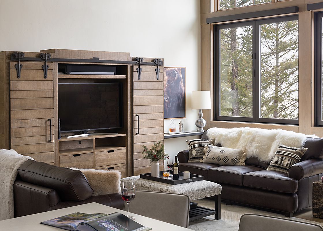 A rustic, cozy living room at The Nest in Jackson Hole with leather sofas, faux-fur throws, a large wooden entertainment center, and scenic forest views.