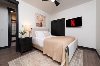 A cozy bedroom with a queen-sized bed, dark wood nightstand, and a wall-mounted TV displaying the Netflix logo.