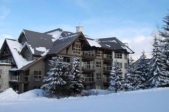A large, multi-story building, Aspens on Blackcomb Unit 113, is nestled among snow-covered evergreen trees with a vast snowfield in the foreground under a clear blue sky.