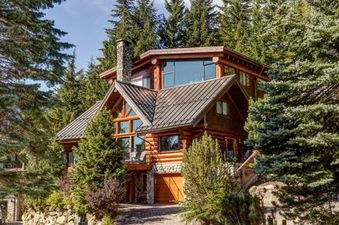 The Snowridge Chalet 7 is a luxurious log cabin-style home nestled among lush evergreen trees, featuring a prominent stone chimney, large windows, and a multi-pitched metal roof.