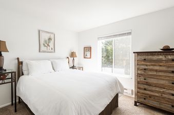A serene bedroom at Snowcrest 300, featuring a bed with white linens, rustic wooden furniture, and a window with a view of greenery.