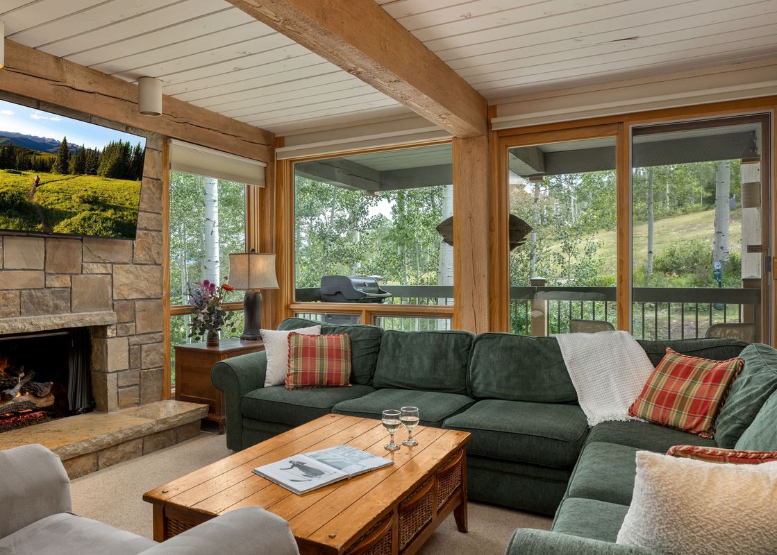 A cozy living room with a green sectional sofa, a stone fireplace, and large windows looking out onto a lush forest.