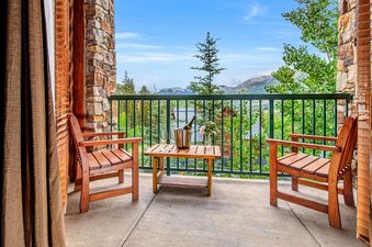 A private balcony with two wooden chairs, a table with champagne and flowers, overlooking a mountain landscape.