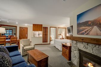A cozy studio apartment at Blackcomb Springs Premium Studio features a living area with a blue sofa, a dining table, a compact kitchen, and a sleeping area with a bed, all illuminated by warm lighting and a fireplace.