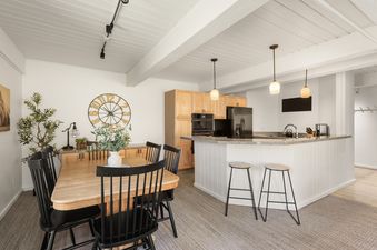 The open-concept dining and kitchen area at Enclave 209 features a wooden dining table with black chairs, a large wall clock, and a modern kitchen with black appliances and a granite-topped island with stools.