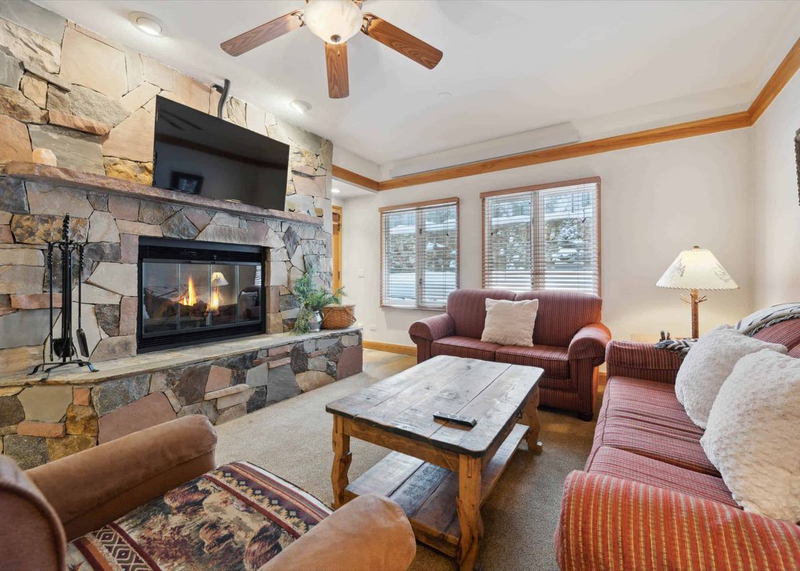 A cozy living room at The Charter F2105 with a stone fireplace, television, two red sofas, a wooden coffee table, and windows showing a snowy exterior.