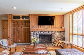Cozy living area in Nez Perce A2 featuring a stone fireplace, wood-paneled walls, and a large window with blinds.