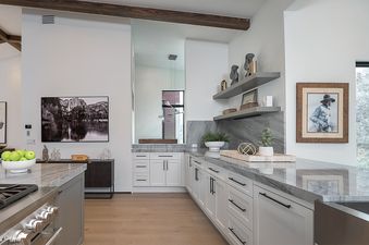 A modern kitchen with white cabinets, marble countertops, and a large island with a gas range. The kitchen is decorated with framed art and various decorative objects.