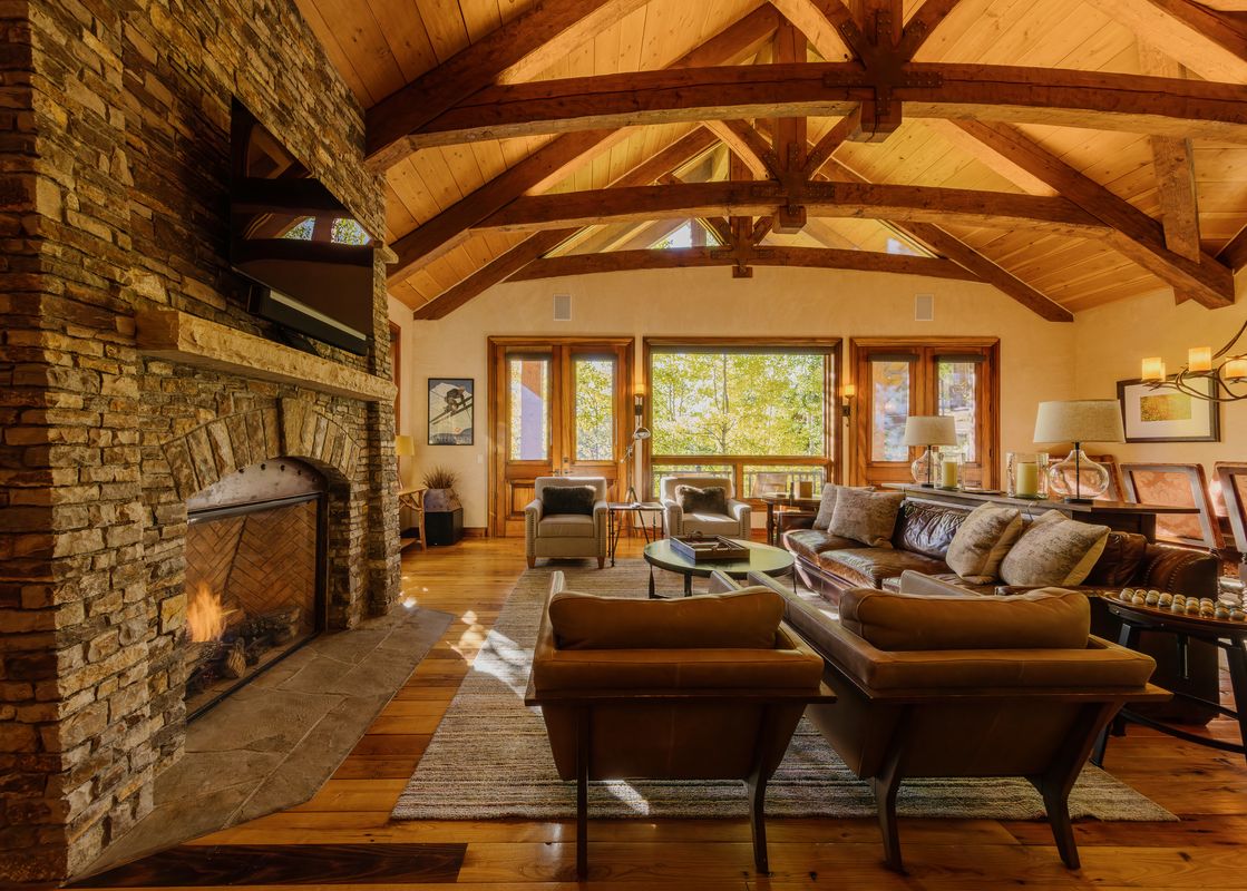 The grand living area at Four Bears Hideaway at See Forever features a stone fireplace, vaulted wooden ceilings, plush leather seating, and large windows with vibrant outdoor views.