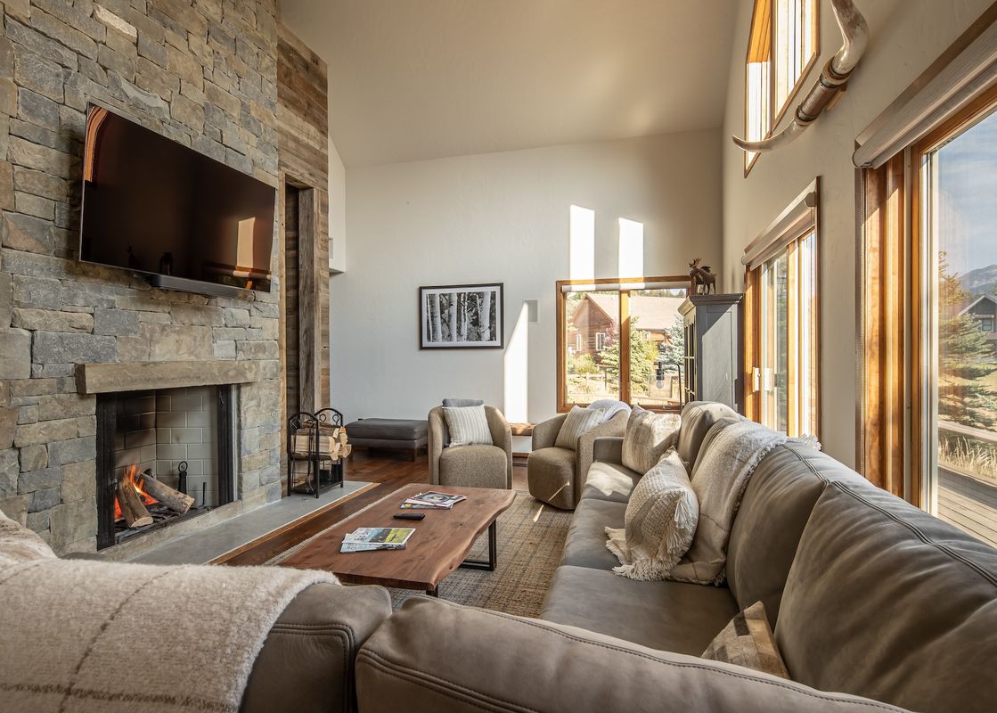 A spacious and inviting living room features a large gray sectional sofa, a live-edge coffee table, and a grand stone fireplace with a mounted television above it, all bathed in natural light from expansive windows.