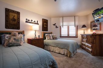 A twin bedroom in Chateau Chamonix CX115 with rustic decor, featuring two beds with green quilts, Western-themed wall art, a window with blinds, and a log-style dresser.